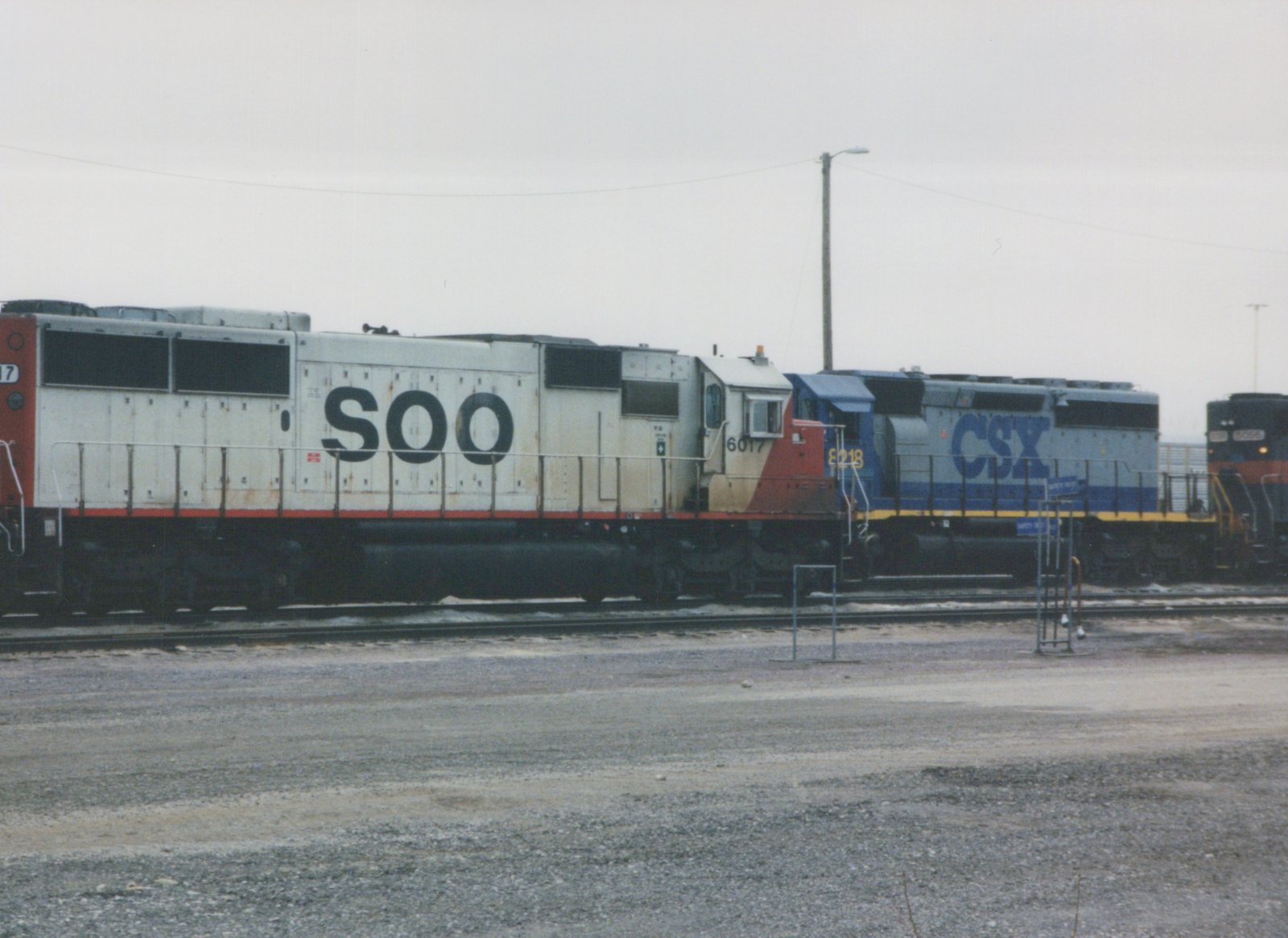 A side view of a white SOO locomotive numbered 6017 parked in a gravel yard next to a blue CSX locomotive and another SOO unit, all under a gray sky.