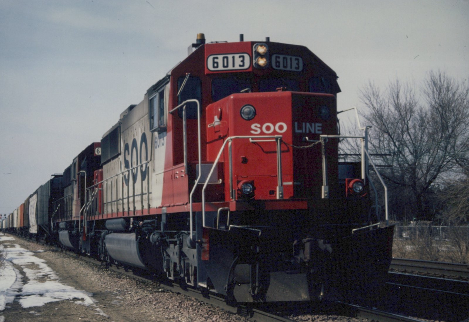 Three-quarter front view of a red and white SOO Line locomotive numbered 6013, pulling freight cars on a snow-dusted track.