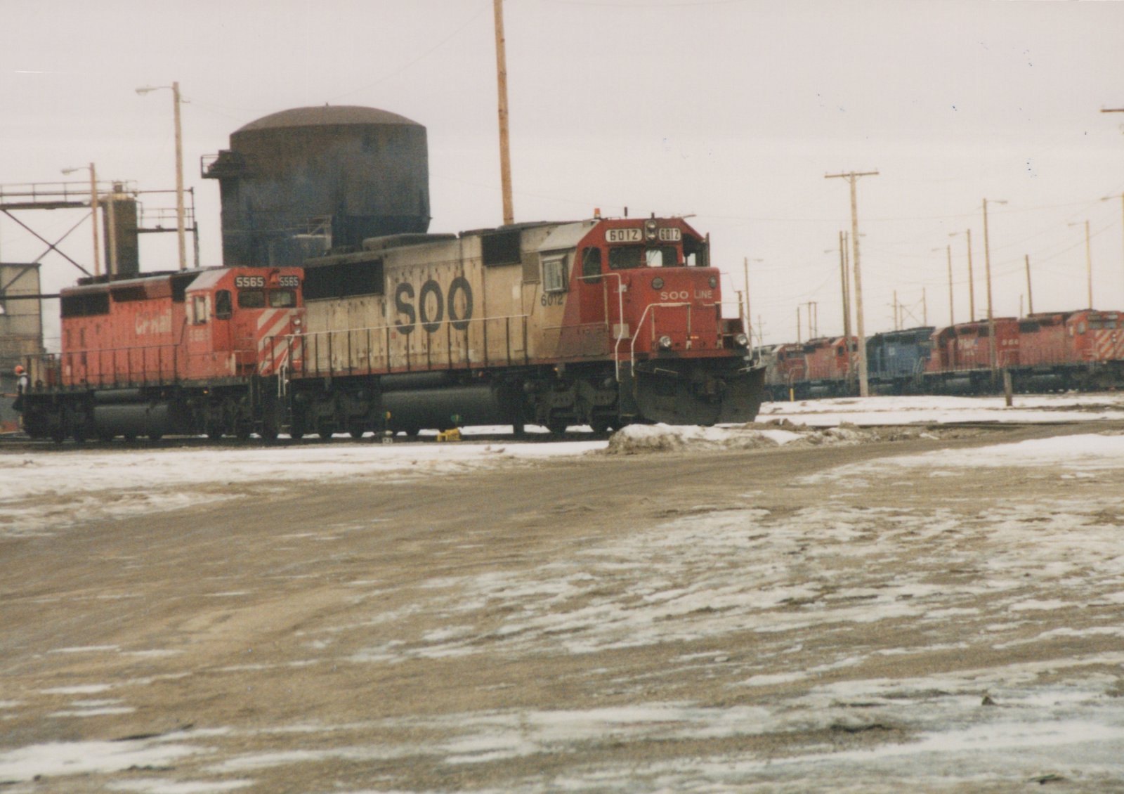 A red SOO Line 6012 diesel locomotive leads a train through a snowy rail yard, with CP Rail locomotives nearby.