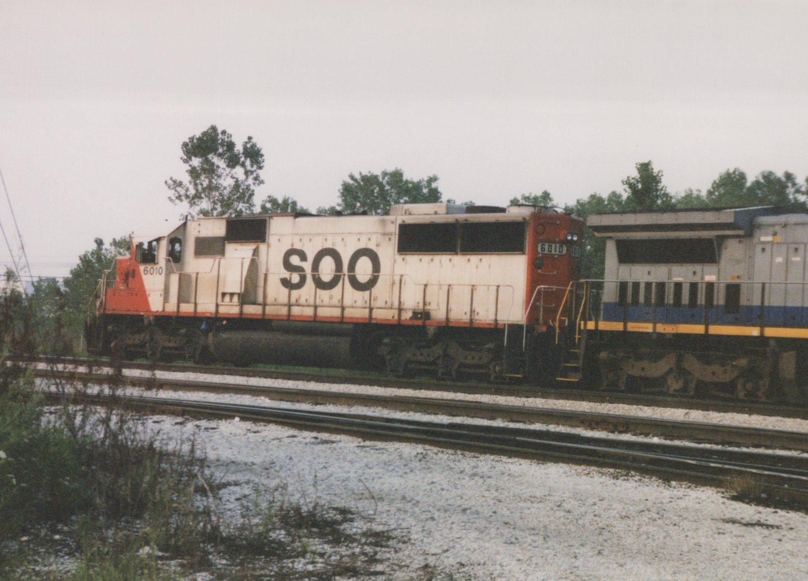 Side view of a white and orange Soo Line diesel locomotive numbered 6010, with an Amtrak blue and silver locomotive behind it, on parallel railroad tracks under an overcast sky.