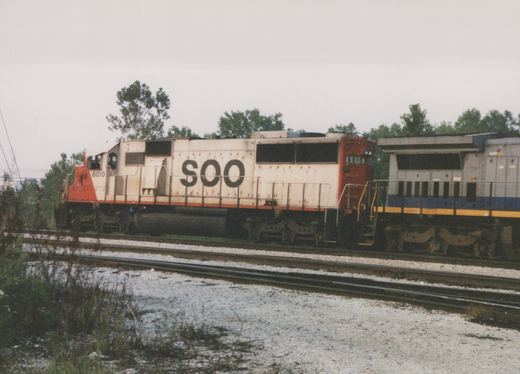 Side view of a white and orange Soo Line diesel locomotive numbered 6010, with an Amtrak blue and silver locomotive behind it, on parallel railroad tracks under an overcast sky.