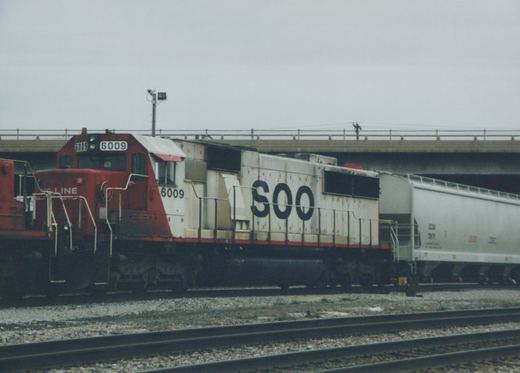 A Soo Line SD40-2 diesel locomotive, number 6009, with red and white paint, is parked on tracks under a concrete overpass. A freight car is visible behind it.