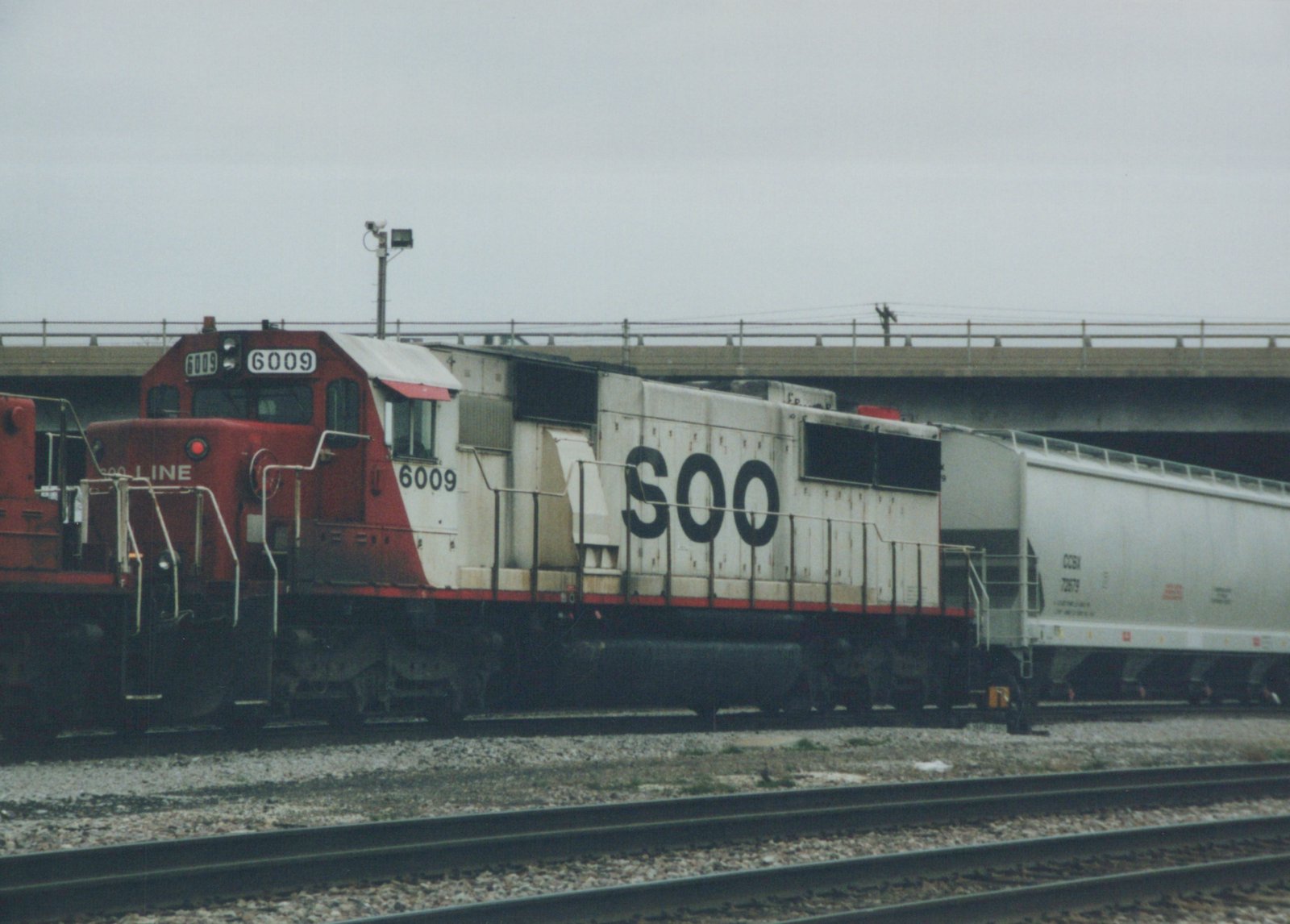 A Soo Line SD40-2 diesel locomotive, number 6009, with red and white paint, is parked on tracks under a concrete overpass. A freight car is visible behind it.