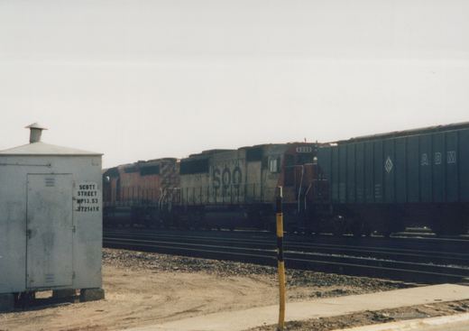 A side view of a grey and red SOO Line locomotive numbered 6008, pulling a dark freight car, stopped on multiple tracks next to a utility box labeled Scott Street under an overcast sky.