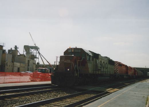 A SOO Line diesel locomotive numbered 6004, leading a train past industrial buildings and a construction vehicle under an overcast sky.
