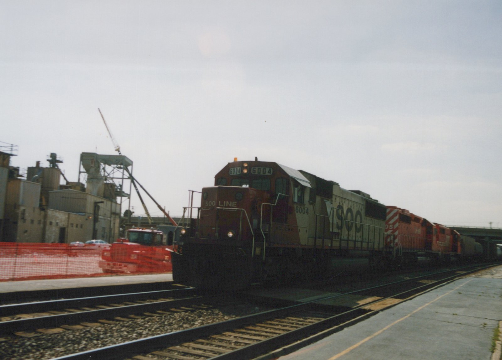 A SOO Line diesel locomotive numbered 6004, leading a train past industrial buildings and a construction vehicle under an overcast sky.