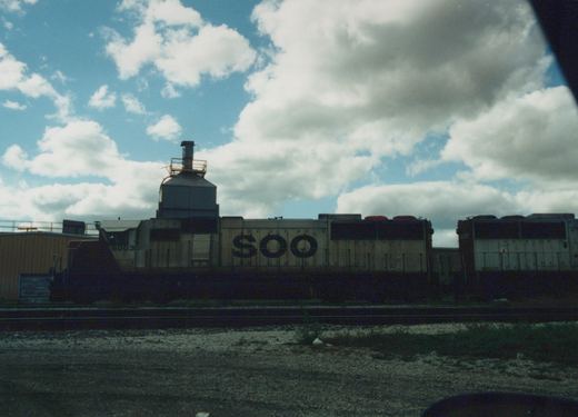 A side view of two SOO Line diesel locomotives, numbered 6003 and 6024, parked on parallel tracks in a gravel yard under a cloudy sky.
