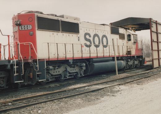 A white and red diesel locomotive with 'SOO' on the side and the number 6001 is on railroad tracks beside a loading dock.