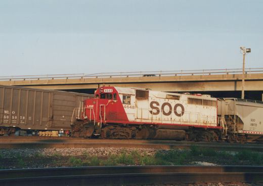 A red and white Soo Line diesel locomotive pulls freight cars under a concrete highway overpass on a clear day.