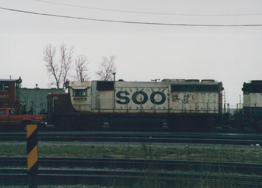 Side view of a white and black Soo Line locomotive number 4603 in a rail yard, with other rail equipment and a red truck in the background, under a gray sky with bare trees.
