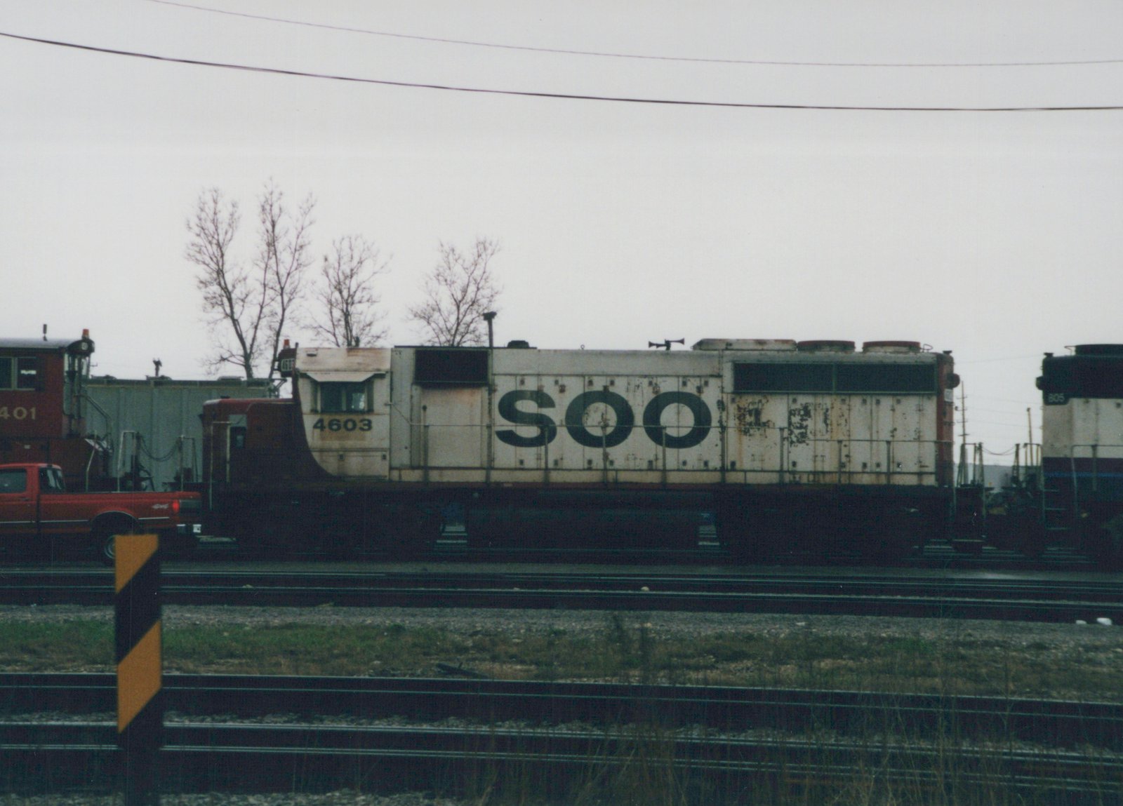 Side view of a white and black Soo Line locomotive number 4603 in a rail yard, with other rail equipment and a red truck in the background, under a gray sky with bare trees.