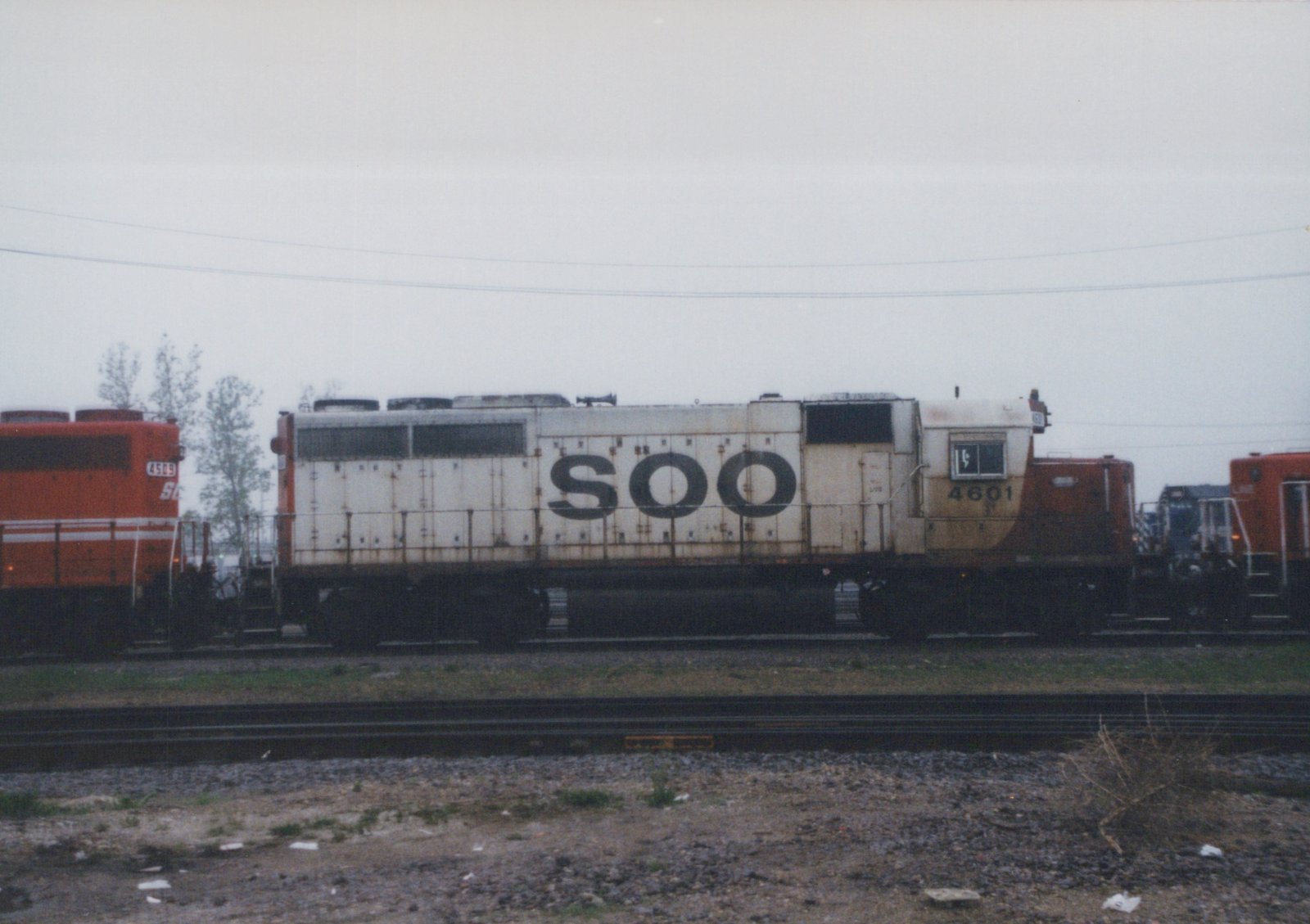 Side view of a white SOO 4601 locomotive with black lettering, parked on tracks next to an orange SOO 4509 locomotive, in a gravel yard under a gray sky.