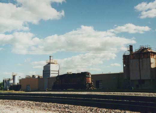 A dark-colored diesel locomotive with the number 4517 and 'SOO' lettering sits on tracks beside industrial buildings under a partly cloudy sky.