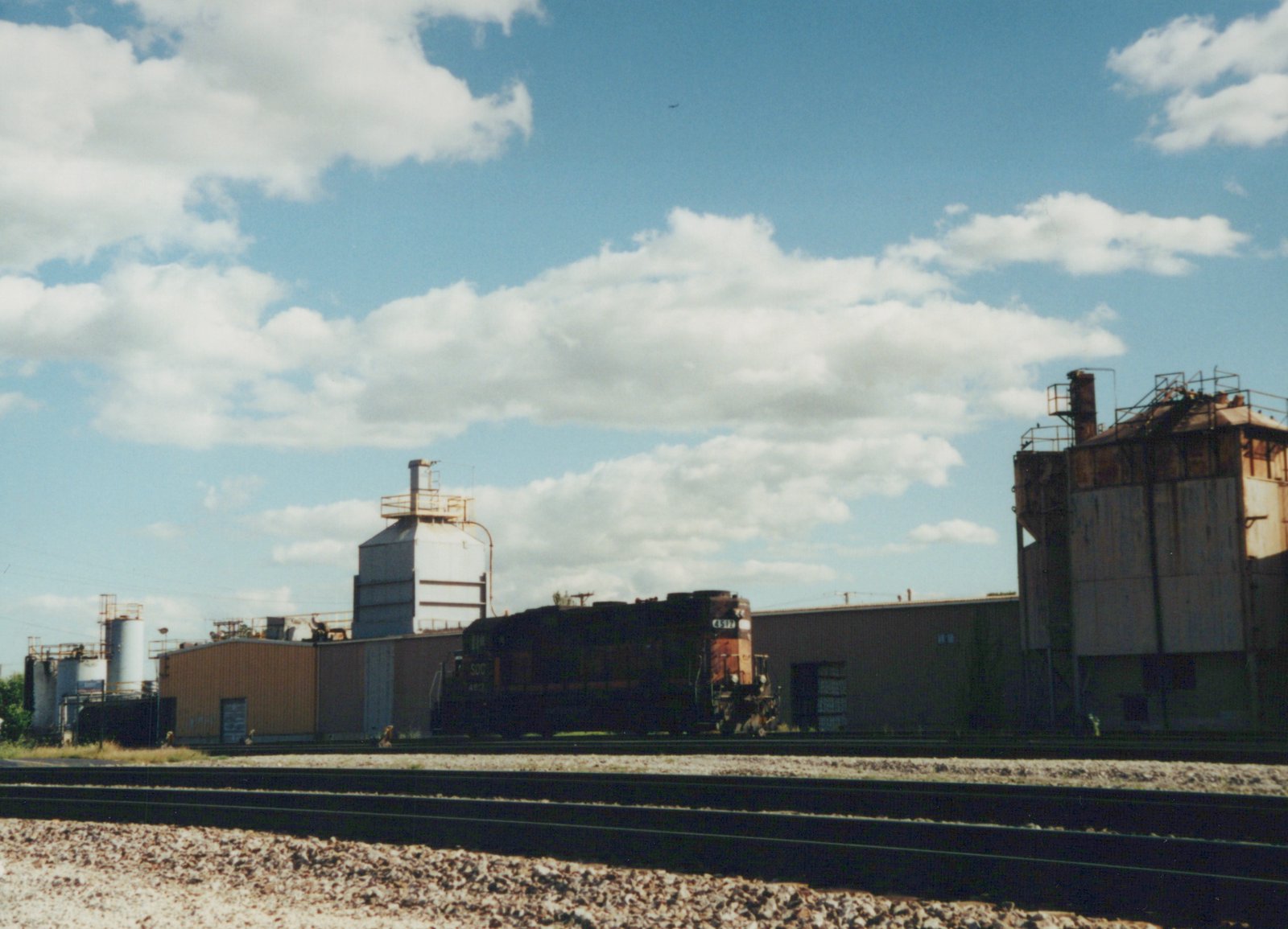 A dark-colored diesel locomotive with the number 4517 and 'SOO' lettering sits on tracks beside industrial buildings under a partly cloudy sky.