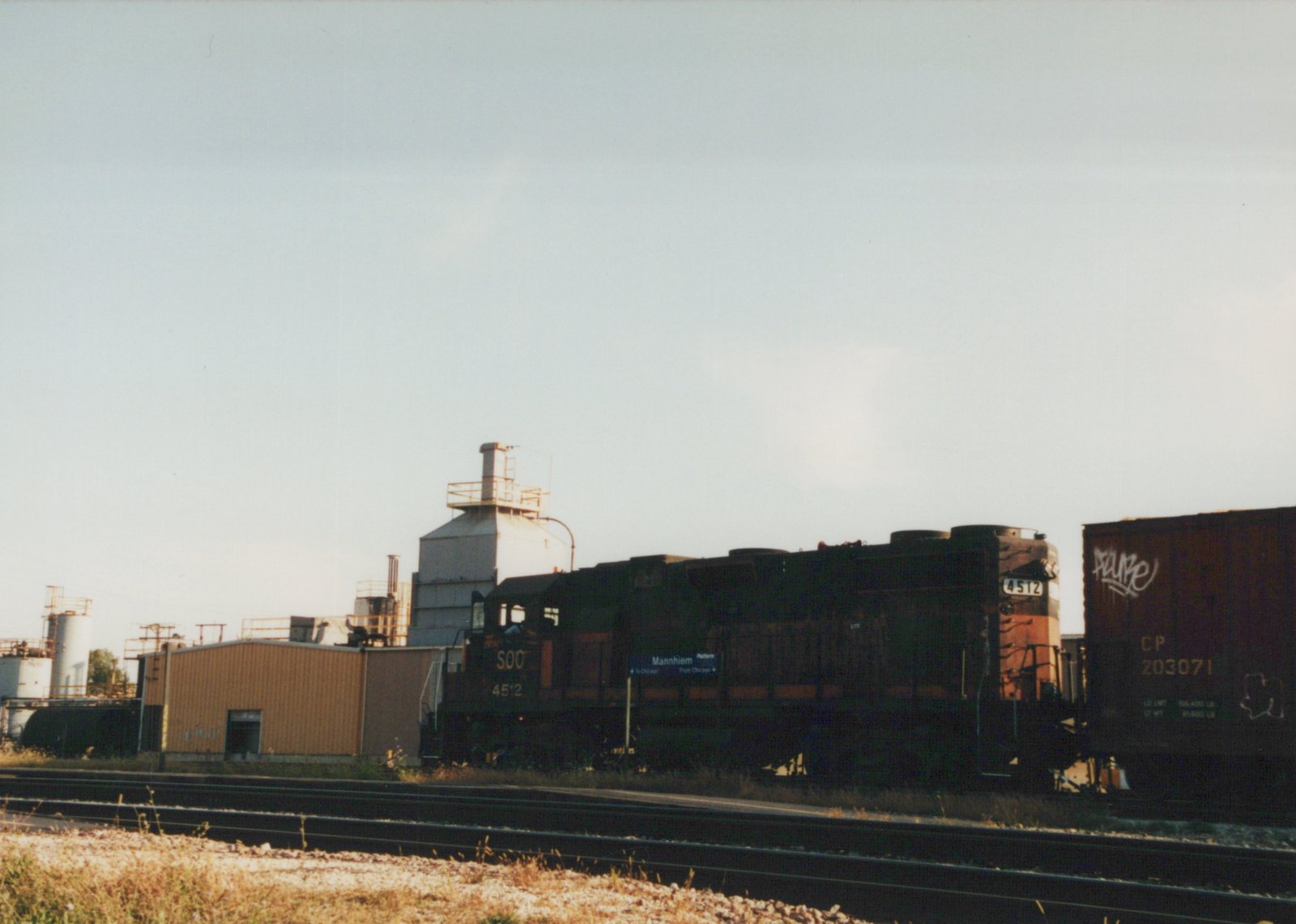 A side view of a dark green and orange Soo Line locomotive numbered 4512 on a set of tracks, pulling a dark brown Canadian Pacific boxcar, with industrial buildings and a 'Mannhiem' station sign in the background.
