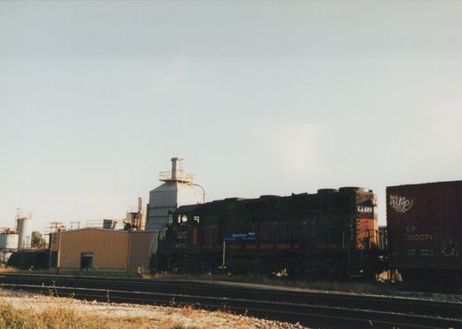 A side view of a dark green and orange Soo Line locomotive numbered 4512 on a set of tracks, pulling a dark brown Canadian Pacific boxcar, with industrial buildings and a 'Mannhiem' station sign in the background.