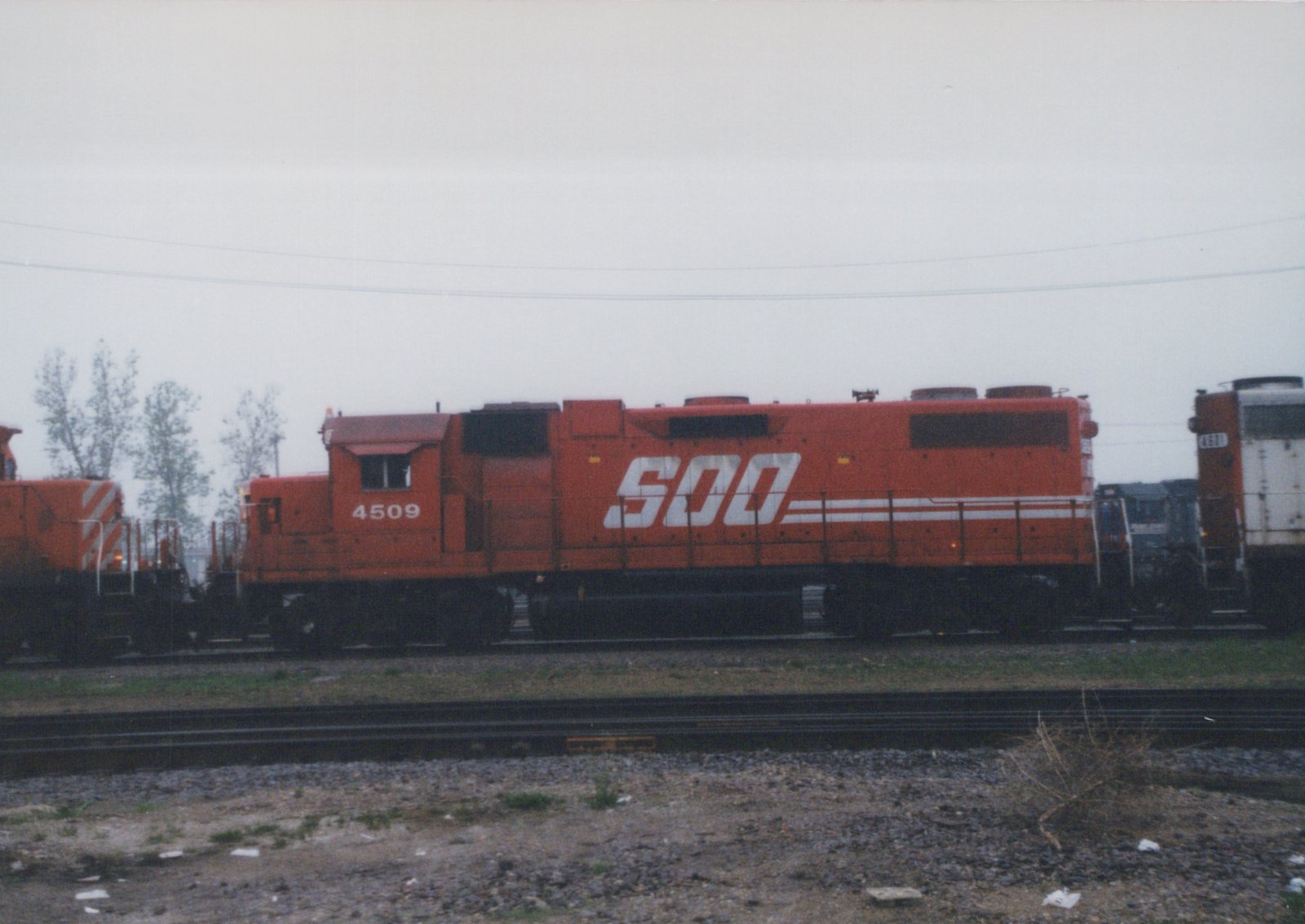An orange SOO Line diesel locomotive #4509 on a set of railroad tracks in a yard under an overcast sky, with another locomotive partially visible behind it.