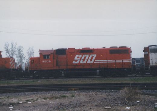 An orange SOO Line diesel locomotive #4509 on a set of railroad tracks in a yard under an overcast sky, with another locomotive partially visible behind it.