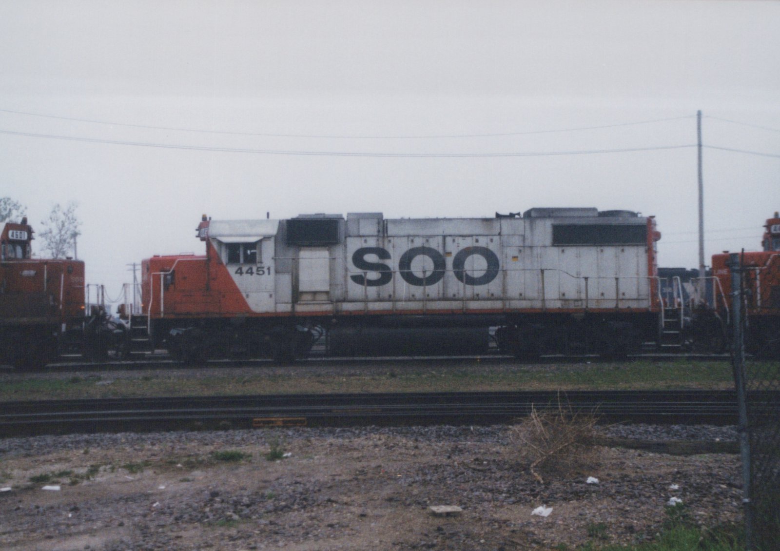 Side view of a white and orange Soo Line diesel locomotive with number 4451, parked in a gravel-yard setting under an overcast sky.