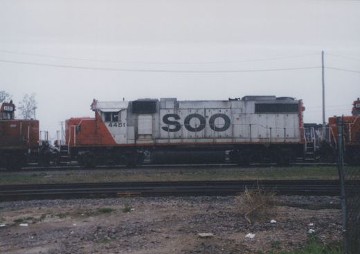 Side view of a white and orange Soo Line diesel locomotive with number 4451, parked in a gravel-yard setting under an overcast sky.