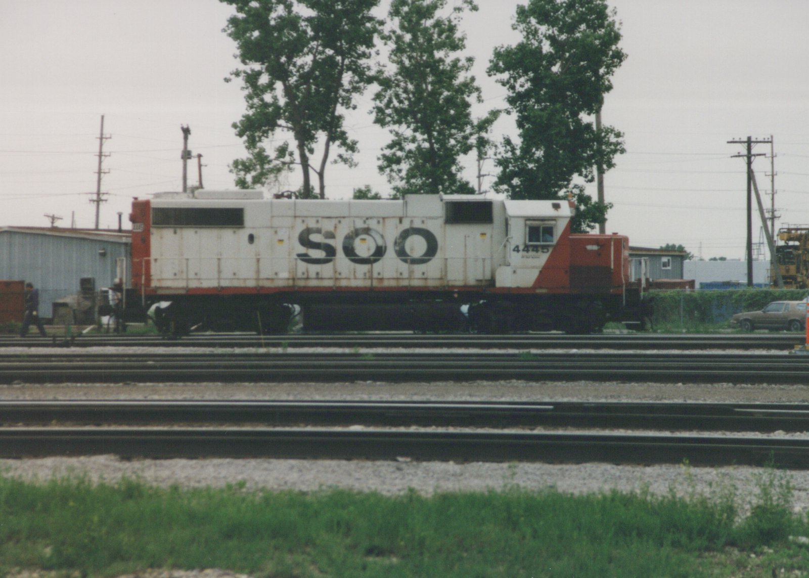 A white and red Soo Line diesel locomotive, number 4445, sits on tracks in a rail yard with trees and a blue building in the background, and one person walking nearby.