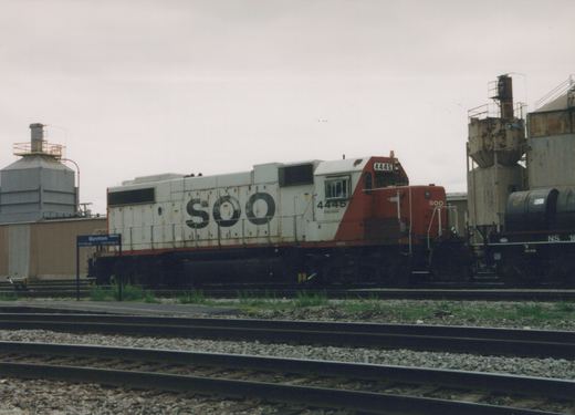 A white and red Soo Line diesel locomotive numbered 4445 is shown on railroad tracks next to a station sign reading 'Mannhiem Platform 2', with an overcast sky and industrial structures in the background.