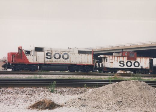 A side view of a white and red SOO Line locomotive with number 4443, coupled to a white and red SOO Line caboose with number 17, on railroad tracks under a concrete overpass, with gravel and weeds in the foreground.