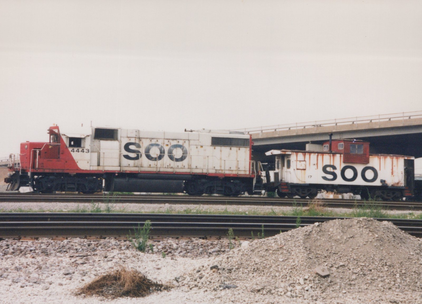 A side view of a white and red SOO Line locomotive with number 4443, coupled to a white and red SOO Line caboose with number 17, on railroad tracks under a concrete overpass, with gravel and weeds in the foreground.
