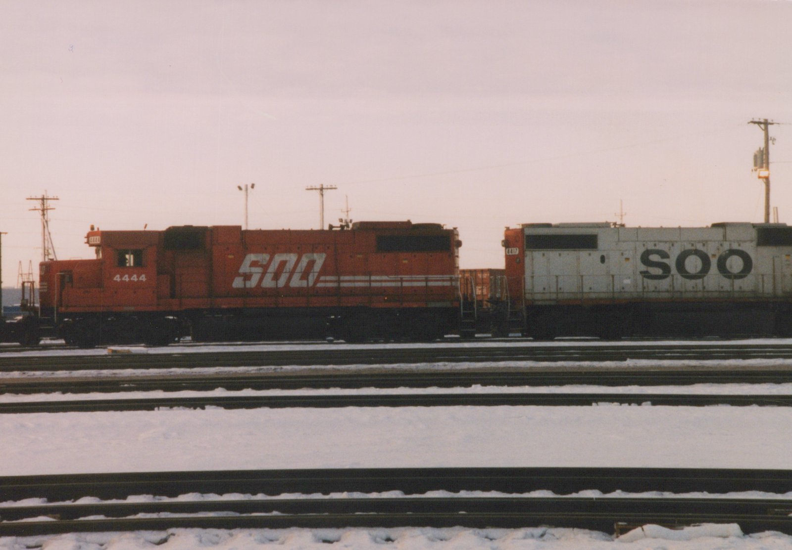 Two SOO Line diesel locomotives, one orange and one white, sit side-by-side on snowy tracks in a rail yard.