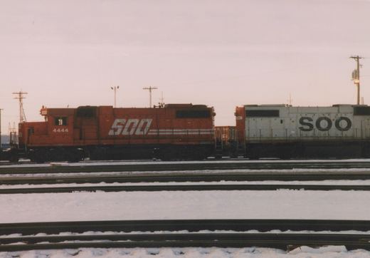 Two SOO Line diesel locomotives, one orange and one white, sit side-by-side on snowy tracks in a rail yard.