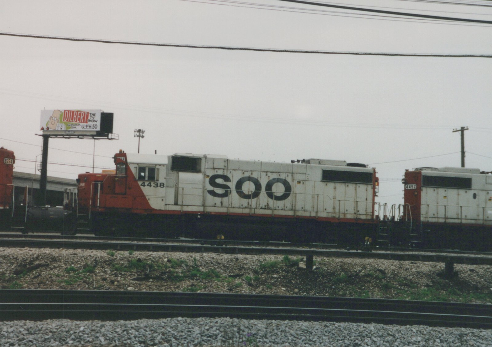 Two SOO Line diesel locomotives, numbered 4438 and 4452, are parked side by side on a railroad track; a billboard for the 'Dilbert' TV show is visible in the background under a gray sky.