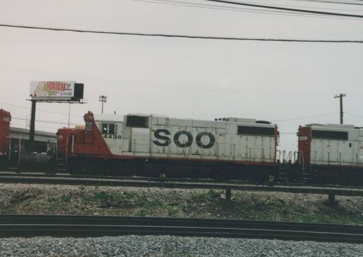 Two SOO Line diesel locomotives, numbered 4438 and 4452, are parked side by side on a railroad track; a billboard for the 'Dilbert' TV show is visible in the background under a gray sky.
