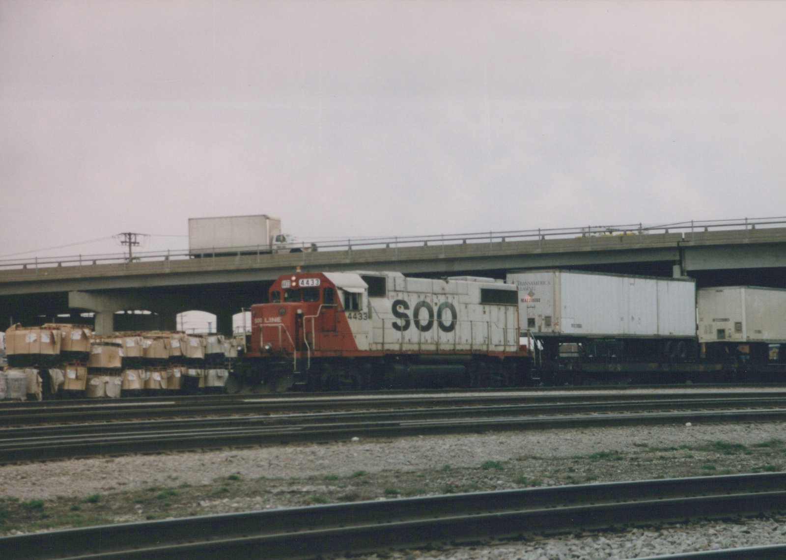 A red and white SOO Line diesel locomotive numbered 4433 pulls a train with trailers on flatcars under a concrete highway bridge on an overcast day.