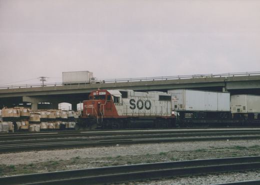 A red and white SOO Line diesel locomotive numbered 4433 pulls a train with trailers on flatcars under a concrete highway bridge on an overcast day.