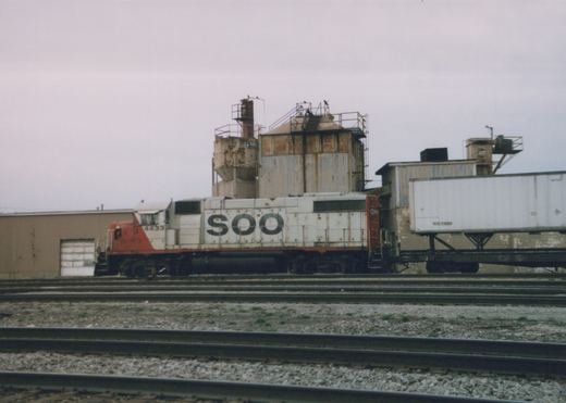 A white and red SOO Line diesel locomotive numbered 4433 sits on tracks in a yard with a white trailer on a flatcar behind it, under an overcast sky.