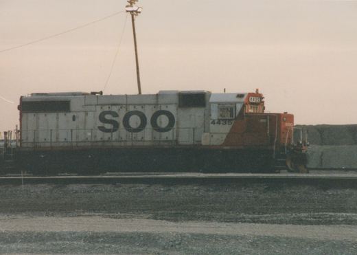 A white and orange SOO Line diesel locomotive, number 4435, parked on a set of tracks in a gravel yard under a cloudy sky.