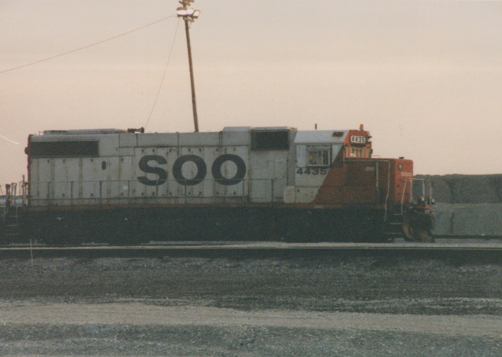 A white and orange SOO Line diesel locomotive, number 4435, parked on a set of tracks in a gravel yard under a cloudy sky.