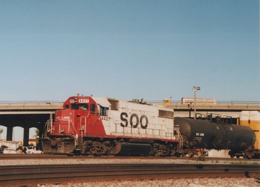 A red and white SOO Line diesel locomotive pulling a black tank car, with a concrete bridge overhead and a white car parked nearby.