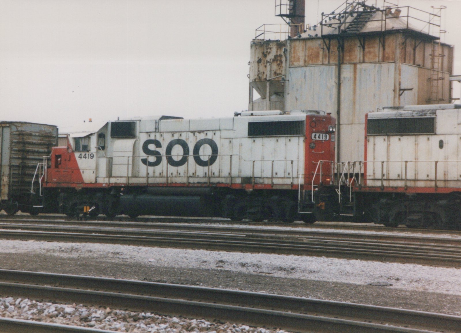 A white and red Soo Line locomotive numbered 4419 sits on tracks in a snow-dusted yard with an industrial building in the background.