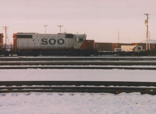 A side view of a white and orange SOO Line diesel locomotive numbered 4417, standing on tracks in a snowy railyard with multiple parallel tracks and freight containers in the background under an overcast sky.