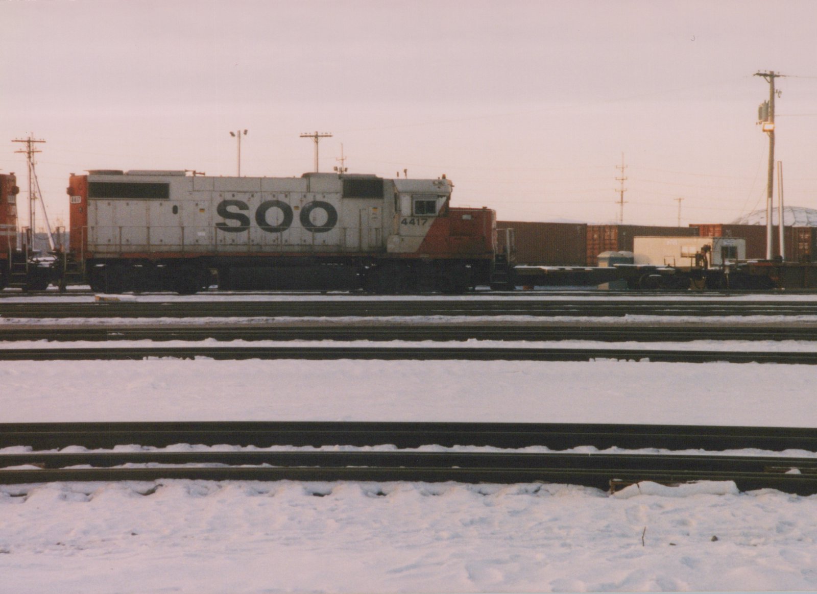 A side view of a white and orange SOO Line diesel locomotive numbered 4417, standing on tracks in a snowy railyard with multiple parallel tracks and freight containers in the background under an overcast sky.