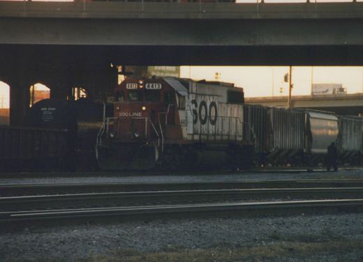 An EMD SD40-2 diesel locomotive painted in SOO Line maroon and white livery, numbered 4413, is traveling on a railroad main line under a concrete overpass during dusk. Several freight cars and a lone person are visible in the background.