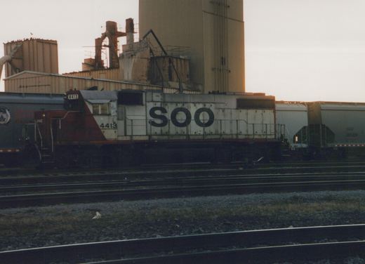 A SOO Line locomotive numbered 4413 is parked on a set of tracks next to freight cars in a rail yard, with industrial buildings in the background at dusk.