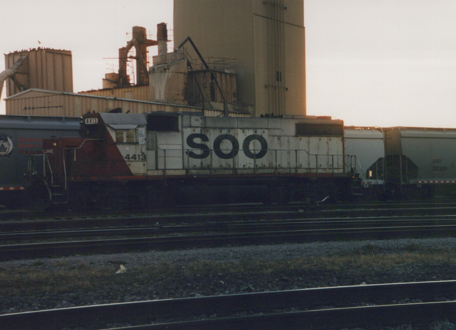 A SOO Line locomotive numbered 4413 is parked on a set of tracks next to freight cars in a rail yard, with industrial buildings in the background at dusk.