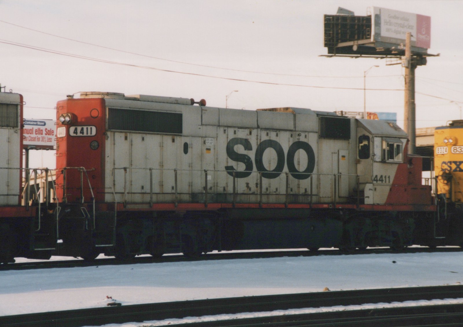 Side view of a Soo Line diesel locomotive numbered 4411, painted white with red ends and black SOO lettering, standing on tracks beside snow-covered ground with an orange CSX locomotive in the background.