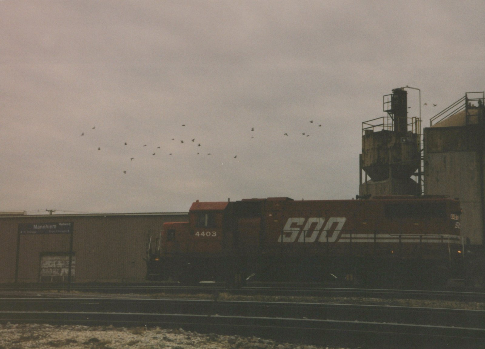 A side view of a red SOO Line diesel locomotive, number 4403, stopped at a station platform under an overcast sky with birds flying overhead.
