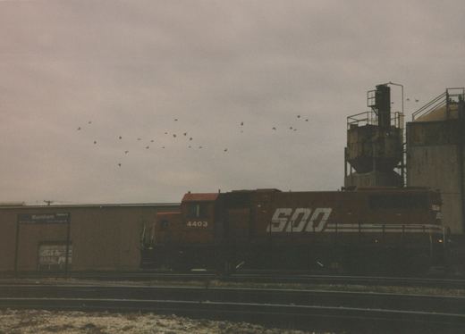 A side view of a red SOO Line diesel locomotive, number 4403, stopped at a station platform under an overcast sky with birds flying overhead.