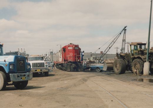 A red Soo Line locomotive with the number 2410 is in a rail yard with a blue Mack truck, a white rental truck, and a front-end loader nearby, under a cloudy sky.