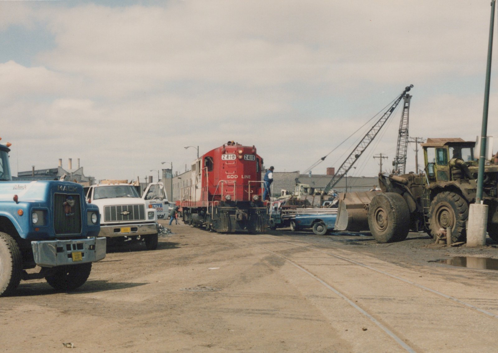 A red Soo Line locomotive with the number 2410 is in a rail yard with a blue Mack truck, a white rental truck, and a front-end loader nearby, under a cloudy sky.