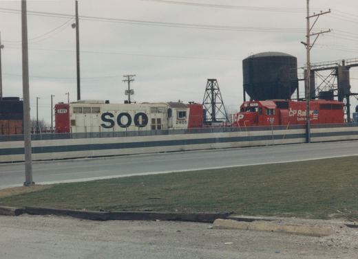 Two diesel locomotives, one white with SOO lettering and the other red with CP Rail lettering, are on a railroad track beside a road, with industrial structures in the background.